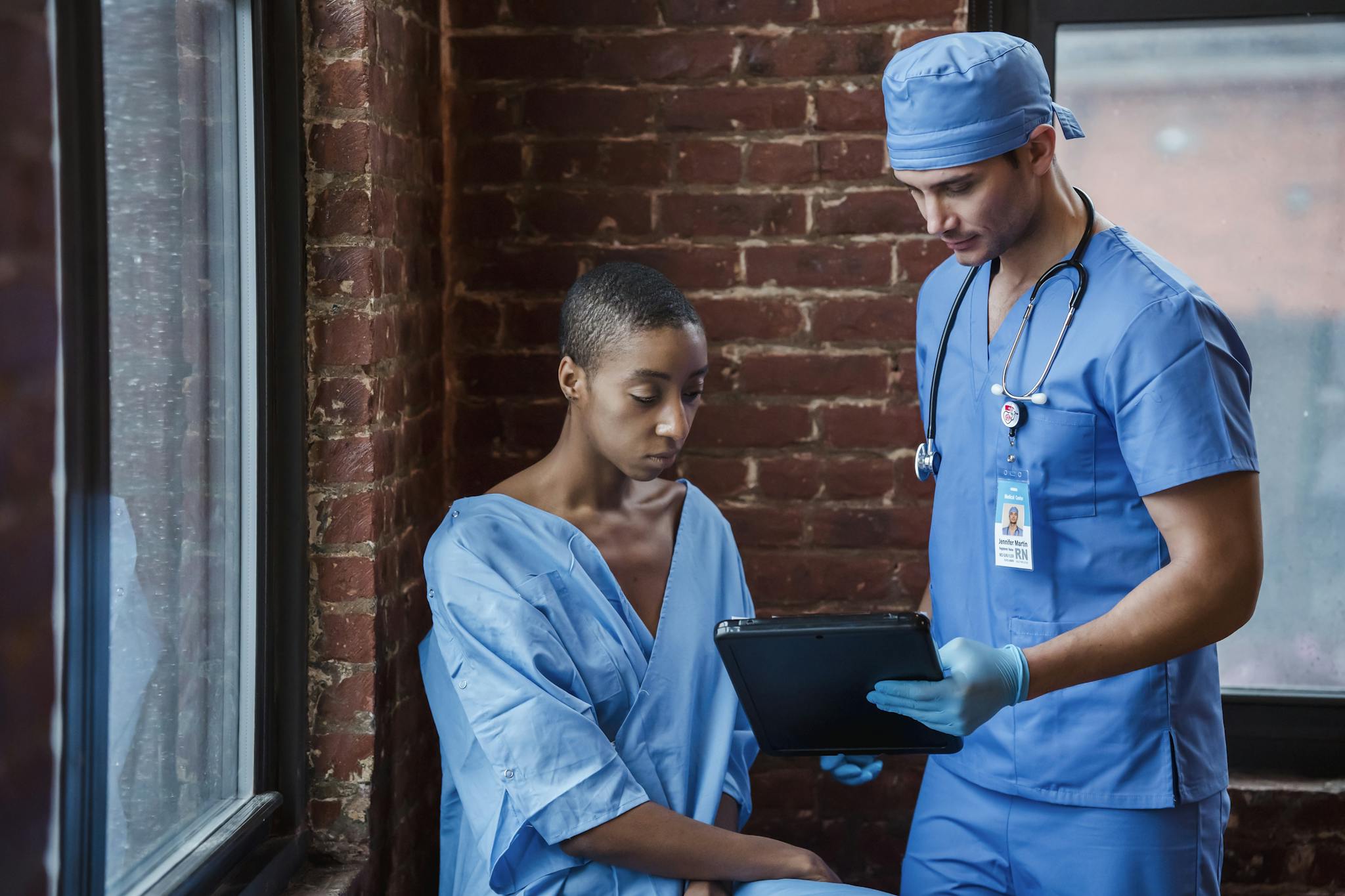 Thoughtful doctor in uniform and gloves showing diagnosis to African American female patient with short hair in blue medical robe in hallway of hospital in daytime